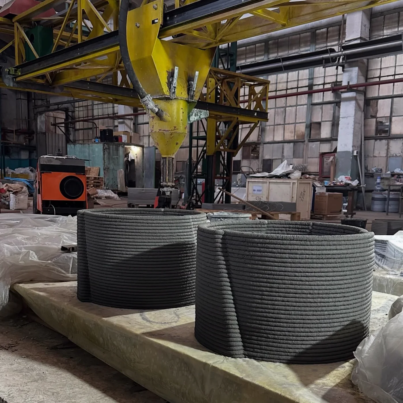 Two freshly printed curved cement-based vessels beneath an industrial extrusion gantry